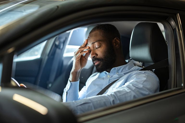 A man inside a vehicle appears tired and stressed, with his hand on his head. Reflects fatigue, busy schedules, and the challenges of everyday transportation.